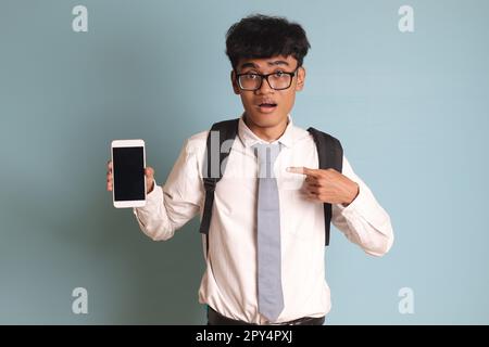 Indonesische Oberschülerin trägt weiße Hemduniform mit grauer Krawatte und präsentiert ein Mobiltelefon mit leerem Bildschirm, während sie auf seine zeigt Stockfoto