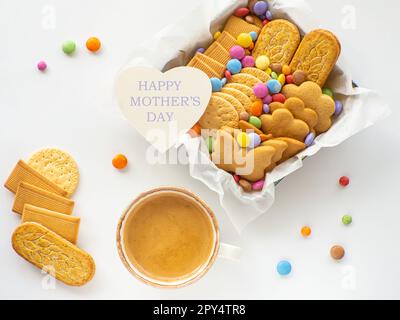 Verschiedene Kekse und Süßigkeiten im Korb mit Happy Mütters Day Note. Handgemachtes Pflegepaket und Tasse Kaffee, Geschenkbox mit Süßigkeiten, Dragee und Keksen Stockfoto