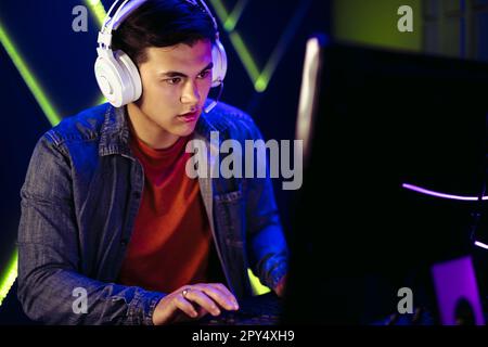 Männlicher Gamer, der mit der Online-Gaming-Welt interagiert. Mit einem sicher aufgesetzten Headset kann er sich voll und ganz auf seinen PC-Bildschirm konzentrieren, während er die Action A im Auge behält Stockfoto