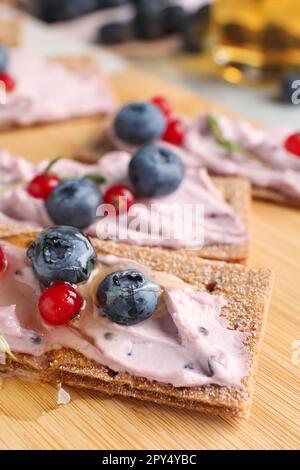 Leckere Cracker-Sandwiches mit Frischkäse, Blaubeeren, roten Johannisbeeren und Honig auf Holzbrett, Nahaufnahme Stockfoto