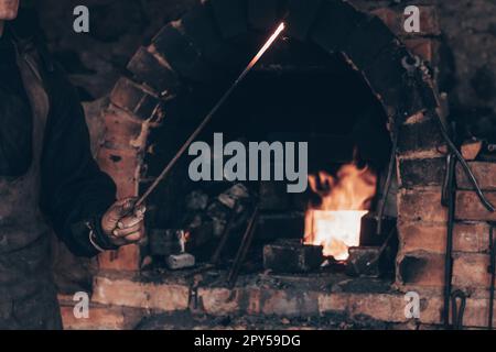 Der Schmied hält ein glühendes geschmolzenes Schwert in der Nähe des Ziegelofens in Schmiede. Farrier in dreckiger Arbeitskleidung, Basteleisen. Stockfoto