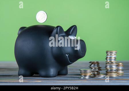 Seitenansicht des schwarzen Sparschweins in der Nähe von Münzstapeln auf dem Tisch auf grünem Hintergrund. Silbermünze, die in die Geldbox fällt. Stockfoto