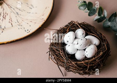 Osterzusammensetzung von Wachtelfleckeneiern in Nestnähe mit Pflanzenzweigen und Eukalyptusästen auf beigefarbenem Hintergrund. Stockfoto