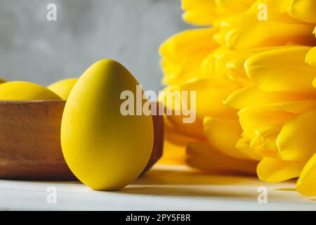 Strauß aus leuchtend gelben Tulpen und Osterei auf weißer Holzoberfläche auf grauem Hintergrund Stockfoto