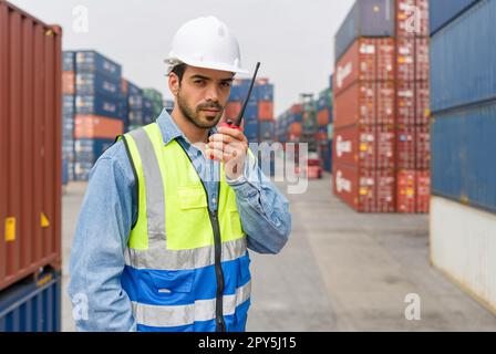 Transportarbeiter mit Sicherheitsweste und Schutzhelm, der mit Walkie-Talkie in der Hand steht. Im Hintergrund gestapelte große Stahlfrachtcontainer. Stockfoto