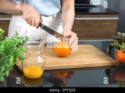 Handgriff mit einem Messer, das Orangenfrüchte auf einem Holzhackbrett schneidet. Das Glas mit gemischtem Fruchtsaft wurde auf die Küchentheke gestellt. Stockfoto