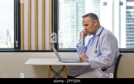 Senior-Professor mit Schnurrbart und Bart liest E-Mails auf einem Laptop. Sie saß am Fenster in der Cafeteria. Vor dem Fenster hat man einen Blick auf die Stadt mit Hochhäusern. Stockfoto