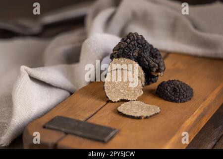 Ganze und geschnittene schwarze Trüffel, Pilze auf Holzbrett auf dunkelbraunem Tisch, Nahaufnahme Stockfoto
