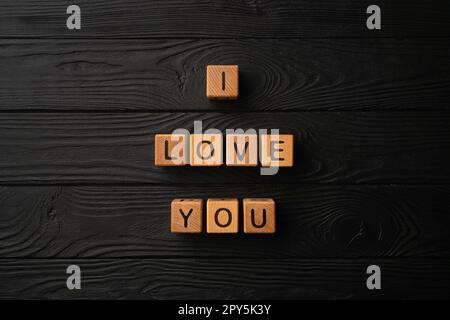 Holzwürfel mit dem Satz "Ich liebe dich" auf schwarzem Hintergrund - ein Holztisch. Liebesworte sind aus Buchstaben aus Holz für deine Freundin, deinen Freund, deine Frau, deinen Ehemann, für den Valentinstag zur Begrüßung einer romantischen Postkarte. Stockfoto
