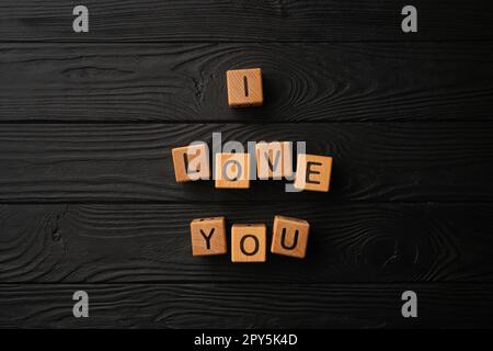 Holzwürfel mit dem Satz "Ich liebe dich" auf schwarzem Hintergrund - ein Holztisch. Liebesworte sind aus Buchstaben aus Holz für deine Freundin, deinen Freund, deine Frau, deinen Ehemann, für den Valentinstag zur Begrüßung einer romantischen Postkarte. Stockfoto