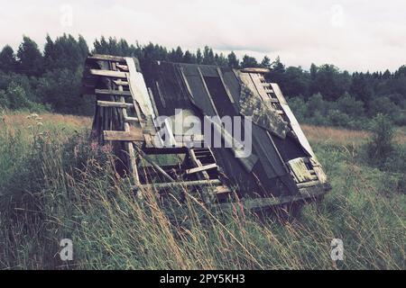 Alte verlassene traurige Quelle oder Haus in Nyrki Dorf, Karelien. Ein einseitiges Holzdach mit verrutschtem Dachmaterial. Ein Feld mit Wiesenblumen und einem Wald in der Ferne. Große Gräser. Sterbende Dörfer. Stockfoto