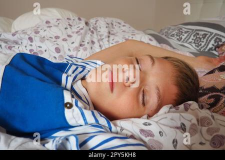 Ein wunderschöner, 10 Jahre alter, weißer Junge mit blondem Haar, gekleidet in gestreiften Pyjamas, schläft auf dem Bett mit einer flauschigen Decke, umarmt ein Kissen. Sanftes Licht der Morgensonne strömt durch das Fenster. Stockfoto