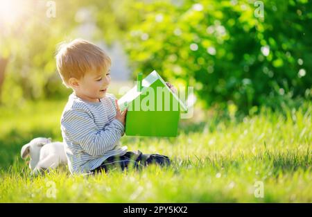 Ein lächelndes Kind, das auf der Frühlingswiese sitzt, mit einem Modell des grünen Hauses in seinen Händen. Stockfoto