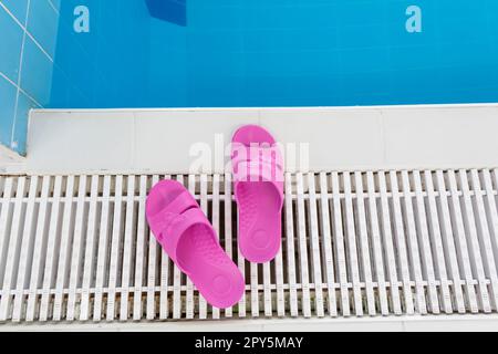 Pinke Hausschuhe stehen am Pool mit blauem Wasser an einem hellen, sonnigen Tag. Stockfoto