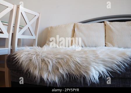 Graues Sofa mit Polsterbezug und weißen, dekorativen Kissen. Weißer Teppich oder Bettüberwurf aus Kunstfell mit langem Stapel. Wohnzimmereinrichtung. Holzstühle. Stockfoto