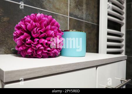 Dekorelemente im Badezimmer. Ein blaues Glas Creme und eine rosafarbene künstliche Doppelblume auf einem Regal. Schwarze Wandfliesen. Bad und WC-Innenausstattung. Heizkörperhandtuchwärmer an der Wand. Stockfoto