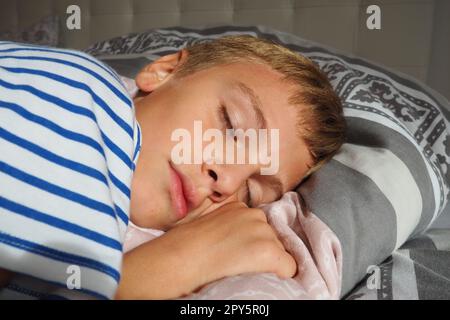 Ein wunderschöner, 10 Jahre alter, weißer Junge mit blondem Haar, gekleidet in gestreiften Pyjamas, schläft auf dem Bett mit einer flauschigen Decke, umarmt ein Kissen. Sanftes Licht der Morgensonne strömt durch das Fenster. Stockfoto