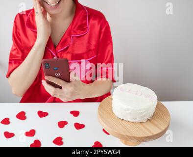 Ein glückliches Mädchen in roter Kleidung hält ein Smartphone in einem Kirschetui mit einem Herzen und ist mit einer SMS von einem geliebten Menschen zufrieden, ein Kuchen und kleine rote Herzen liegen auf dem Tisch. Valentinstag- und romantisches Date-Konzept. Stockfoto