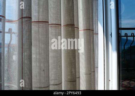 Weiße, grobe Baumwolle- oder Leinenvorhänge aus Gewebungen aus dünnen Fäden. Einfache, kompakte und kostengünstige Wohnungseinrichtung. Fensterdekoration im Zimmer. Roter Streifen. Stockfoto