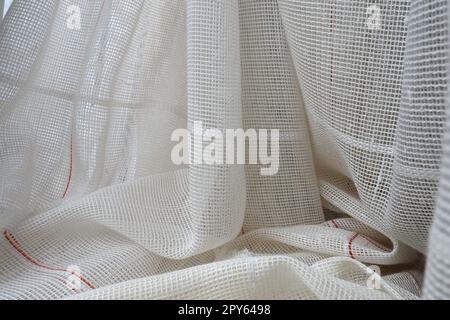 Weiße, grobe Baumwolle- oder Leinenvorhänge aus Gewebungen aus dünnen Fäden. Einfache, kompakte und kostengünstige Wohnungseinrichtung. Fensterdekoration im Zimmer. Roter Streifen. Stockfoto