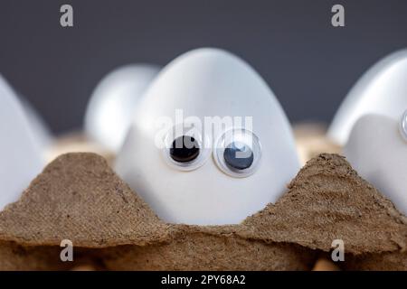 Lustige Gesichter auf weißen Eiern in einer Schachtel mit Bio-Hühnereiern auf dem Küchentisch, die große Animationsaugen zusammenschließen. Humor, Essen und osterferienkonzept. Stockfoto