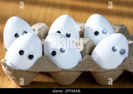 Lustige Gesichter auf weißen Eiern in einer Schachtel mit Bio-Hühnereiern auf dem Küchentisch, die große Animationsaugen zusammenschließen. Humor, Essen und osterferienkonzept. Stockfoto