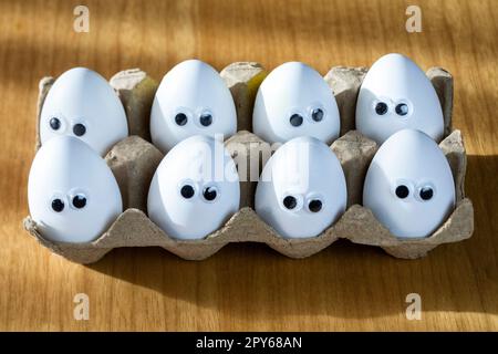 Lustige Gesichter auf weißen Eiern in einer Schachtel mit Bio-Hühnereiern auf dem Küchentisch, die große Animationsaugen zusammenschließen. Humor, Essen und osterferienkonzept. Stockfoto