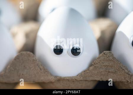 Lustige Gesichter auf weißen Eiern in einer Schachtel mit Bio-Hühnereiern auf dem Küchentisch, die große Animationsaugen zusammenschließen. Humor, Essen und osterferienkonzept. Stockfoto