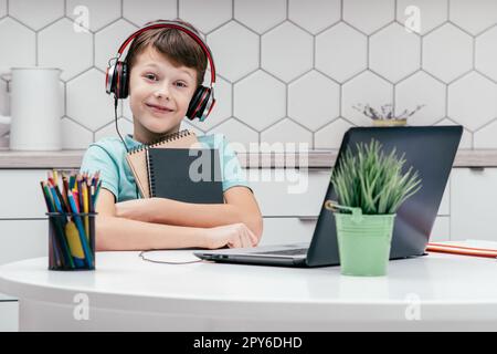 Porträt eines jungen, süßen Jungen, der T-Shirt, Kopfhörer trägt, vor dem Laptop sitzt, kuschelige Notizbücher in der Hand hält. Stockfoto