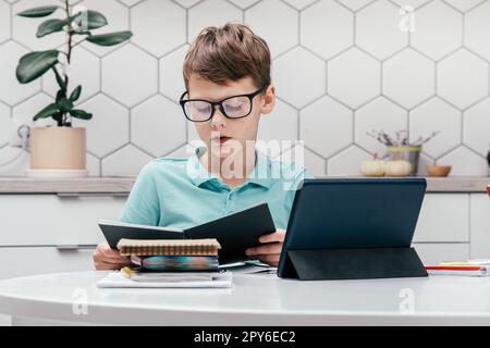 Porträt eines jungen Kindes, der eine Brille trägt, sitzt, sich ein Buch in der Nähe eines Tablets ansieht, Online-Unterricht hat, liest. Stockfoto