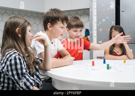 Fröhliche kleine Schulfreunde spielen Brettspiel auf einer Karte mit Würfel und Chips am Tisch, Seitenansicht. Interessantes Tischspiel Stockfoto