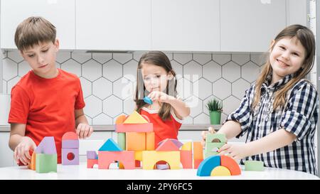 Fröhliche Schulfreunde spielen Baumeister auf dem Küchentisch und sammeln farbenfrohe Details von lego. Kleine Kinder bauen eine Spielzeugstadt. Stockfoto