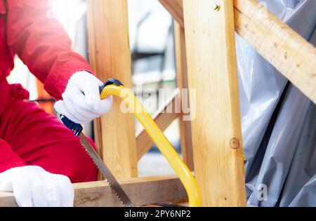 Ein Arbeiter sägt Holz, um eine Kiste für den Transport einer Industriemaschine zu montieren. Eine Frau in rotem Mechaniker-Overall, die eine Bogensäge hält und die Holzbohle einer Kiste schneidet. Konzept Sicherheit am Arbeitsplatz. Stockfoto