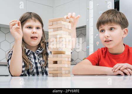 Kinder spielen Jenga, sitzen am Tisch. Junge und Mädchen mit konzentriertem Gesichtsausdruck bauen Turm aus Holzblöcken. Stockfoto