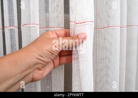 Weiße, grobe Baumwolle- oder Leinenvorhänge aus Gewebungen aus dünnen Fäden. Einfache, kompakte und kostengünstige Wohnungseinrichtung. Fensterdekoration im Zimmer. Die Hand einer Frau streicht den Tüll, zeigt die Qualität. Stockfoto