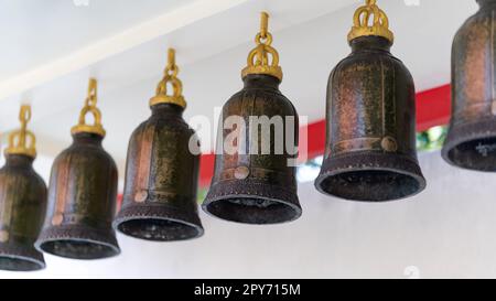 Sechs traditionelle bronzefarbene Glocken hängen eine nach der anderen an eine weiße Wand. Stockfoto