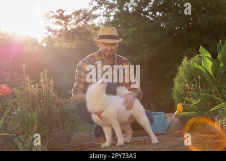 Ein lächelnder Kaukasier in legeren Kleidern, mit einem weißen, samoylierten Welpen. Sonniger Garten im Hintergrund. Das Konzept des Trainingstiers und des glücklichen Hundes Stockfoto