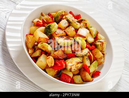 In der Pfanne gebräunte Kartoffeln mit rotem Paprika und ganze Knoblauchzehen in einer weißen Schüssel Stockfoto