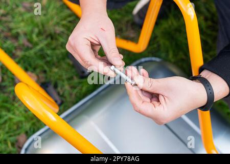 Auf der Straße steht eine Schubkarre aus Metall, ein Mann dreht eine neue Schubkarre für die Arbeit, verdreht Bolzen und Muttern. Stockfoto