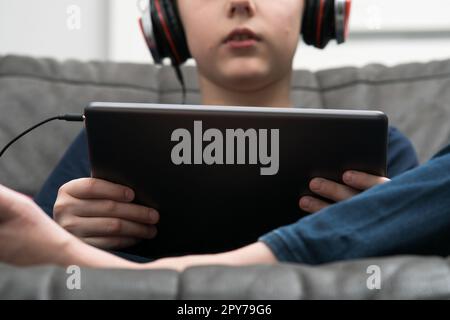 Verkürzter Junge im Mittelteil in kabelgebundenen Kopfhörern, verbunden mit einem digitalen Tablet, sitzt auf dem Sofa. Online-Kurs, Heimunterricht Stockfoto