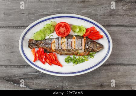 Tischplatte, gegrillte Forelle auf dem ovalen Schild, Orangenscheiben, Tomaten, Paprika, Gurken und Frühlingszwiebeln. Stockfoto