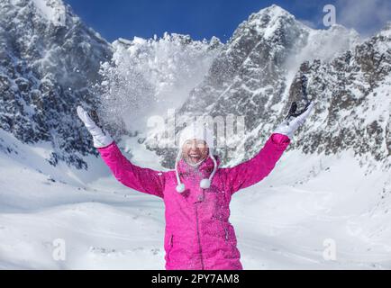 Junge Frau in pinkfarbener Skijacke, Handschuhen und Wintermütze, lächelt, wirft Schnee in die Luft, Sonne scheint auf den Berg hinter ihr. Skigebiet Skalnate Pleso, Slowakei. Stockfoto