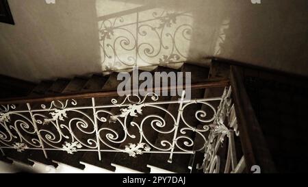 Geheimnisvolle Treppe mit einem geschmiedeten Metallgeländer in der Nacht in der Dunkelheit mit schwachem Licht. Reflexion von Curlicues und Fälschungsmustern. Geländer und Stufen. Treppe im Museum Sremska Mitrovica Serbien Stockfoto