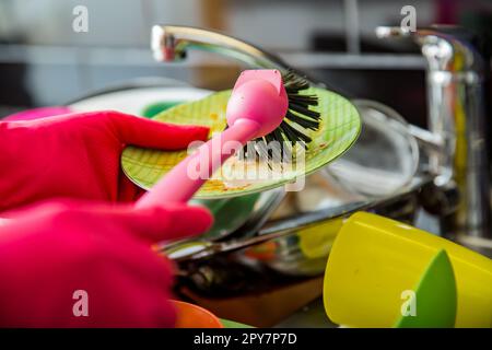 Das Waschbecken war voller schmutzigem Geschirr. Eine Frau in rosa Gummihandschuhen, die mit einem Pinsel abspült. Dunkle Arbeitsfläche in moderner Küche. Hausarbeiten Stockfoto
