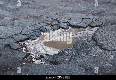 Die befestigte Straßenoberfläche ist beschädigt. Gebrochener Asphalt. Pfütze auf gerissener Straße. Stockfoto