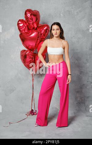 Fröhliche junge Frau in einer breiten, leuchtend pinkfarbenen Hose und weißem Oberteil, die neben Ballons posiert Stockfoto