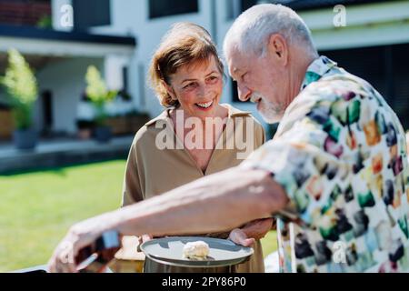Ein älterer Mann grillte draußen im Garten und gab seiner Frau einen Hamburger. Stockfoto