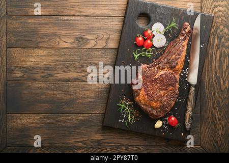 Tomahawk-Steak. Gegrilltes Tomahawk-Rindersteak mit gebackenen Kirschtomaten, Kräutern und Salz auf altem Holzhintergrund. Grillvorgang wird vorbereitet. Draufsicht Stockfoto