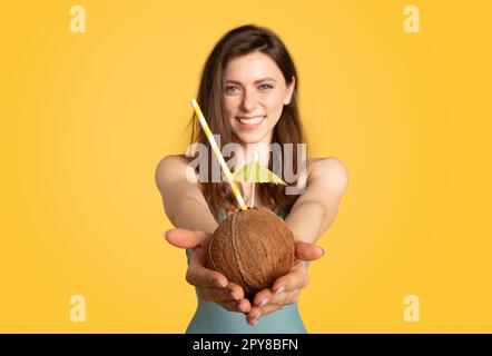 Glückliche wunderschöne Dame im Badeanzug, die vor gelbem Hintergrund steht, mit Kokosnuss in der Hand, selektiv auf frischen Cocktail fokussiert Stockfoto