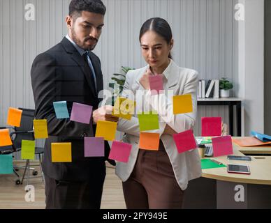 Geschäftsmann in schwarzem Anzug und Arbeitskollegen treffen sich mit Haftnotizen an der Glaswand für neue Ideen. Einsatz agiler Methoden für Unternehmen in einem Tech-Start-up-Büro. Stockfoto